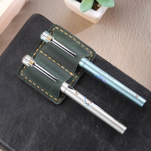 May include: A green leather pen holder with two pens. The holder has two slots for pens and is stitched with yellow thread. One pen is silver and the other is light blue with Chinese characters.