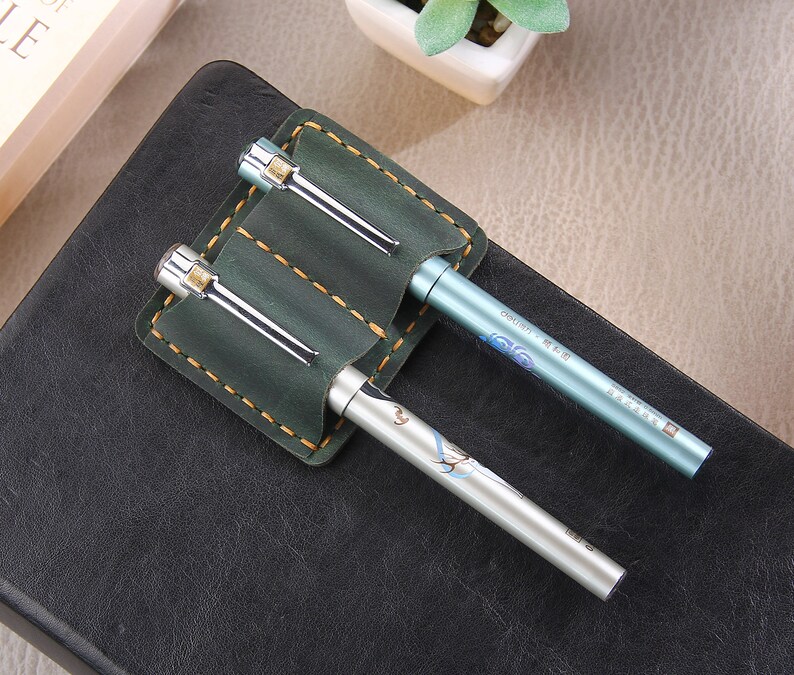 May include: A green leather pen holder with two pens. The holder has two slots for pens and is stitched with yellow thread. One pen is silver and the other is light blue with Chinese characters.