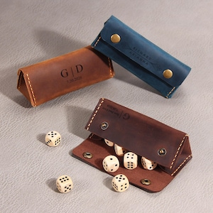 May include: Three leather cases for dice, including one brown case engraved with G|D 7.20.2026, one blue case engraved with RICHARD ARIANNA, and one open dark brown case engraved with Best Dad Ever 7.20.2026, accompanied by several wooden dice with black pips.