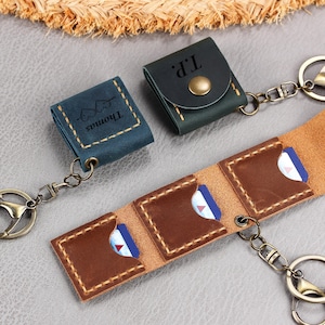 Könnte beinhalten: Drei Schlüsselanhänger-Etuis aus Leder mit Metallbeschlägen. Ein blaues Etui ist mit "Thomas" und einem Herzmotiv graviert. Ein dunkelgrünes Etui ist mit "T.P." graviert und hat einen Messingdruckknopf. Ein braunes Etui ist offen und zeigt drei Fächer.