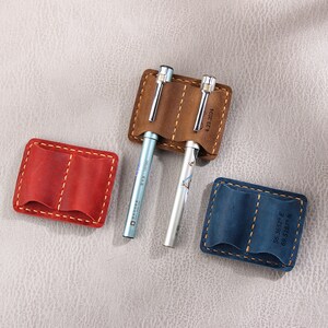 May include: Three leather pen holders with stitched edges. The holders are brown, red, and blue. The brown holder has the date "4.23.2021" printed on it. The blue holder has the coordinates "56.3852&deg; E 69.5187&deg; N" printed on it.