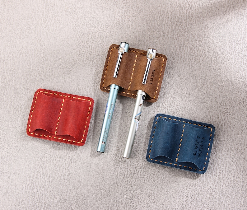 May include: Three leather pen holders with stitched edges. The holders are brown, red, and blue. The brown holder has the date "4.23.2021" printed on it. The blue holder has the coordinates "56.3852&deg; E 69.5187&deg; N" printed on it.