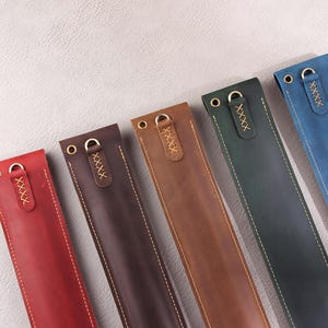 May include: Six leather pen cases in different colors, including red, brown, dark brown, blue, and green. Each case has a gold metal ring and a stitched design.