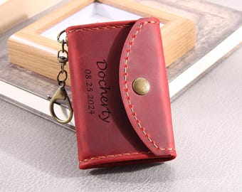 May include: A red leather key pouch featuring a brass snap button closure, visible light-colored stitching, and an attached brass-toned metal keychain. The front is engraved with the text "Docherty" and the date "08.25.2024" in a dark font.
