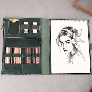 May include: A green leather sketchbook with a zipper closure and a built-in pencil holder. The sketchbook has a drawing of a woman with a flower in her hair on the front.