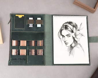 Puede incluir: Un cuaderno de bocetos de cuero verde con cierre de cremallera y un porta lápices incorporado. El cuaderno de bocetos tiene un dibujo de una mujer con una flor en el pelo en la parte delantera.