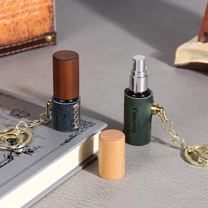 May include: Two portable spray bottles with wooden caps and leather sleeves attached to gold-toned keychains. One sleeve is dark blue with the name Claire, and the other is dark green with the name Cameron. A separate light wooden cap sits nearby.