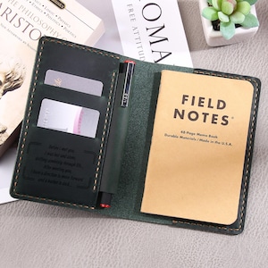 May include: A green leather notebook cover with a pen holder and two card slots. The cover has an engraved quote. The notebook is a Field Notes brand memo book with 48 pages. The notebook is made in the USA.