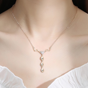 Può includere: Collana in oro con un delicato design a ramo e tre ciondoli a goccia che compongono la parola "MOM".