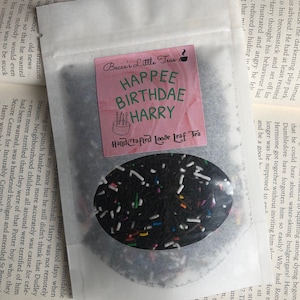 May include: A white paper bag with a pink label that says "Becca's Little Teas" and "HAPPEE BIRTHDAE HARRY" in black text. The label has a hand-drawn image of a birthday cake with candles. The bag contains loose leaf tea with black tea leaves and colourful sprinkles.