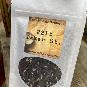 May include: A white paper bag with a label that reads "Becca's Little Boutique 221b Baker St. Handcrafted Loose Leaf Tea". The bag contains loose leaf tea.