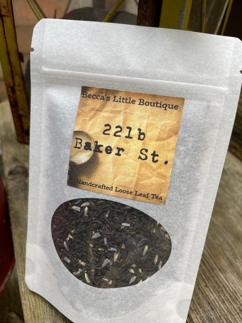May include: A white paper bag with a label that reads "Becca's Little Boutique 221b Baker St. Handcrafted Loose Leaf Tea". The bag contains loose leaf tea.
