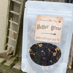 May include: A white resealable pouch with a clear window showing loose leaf tea. The label reads "Becca's Little Teas" and "Butter Brew" with a hand-drawn illustration of a broom. The label also says "Handcrafted Loose Leaf Tea".
