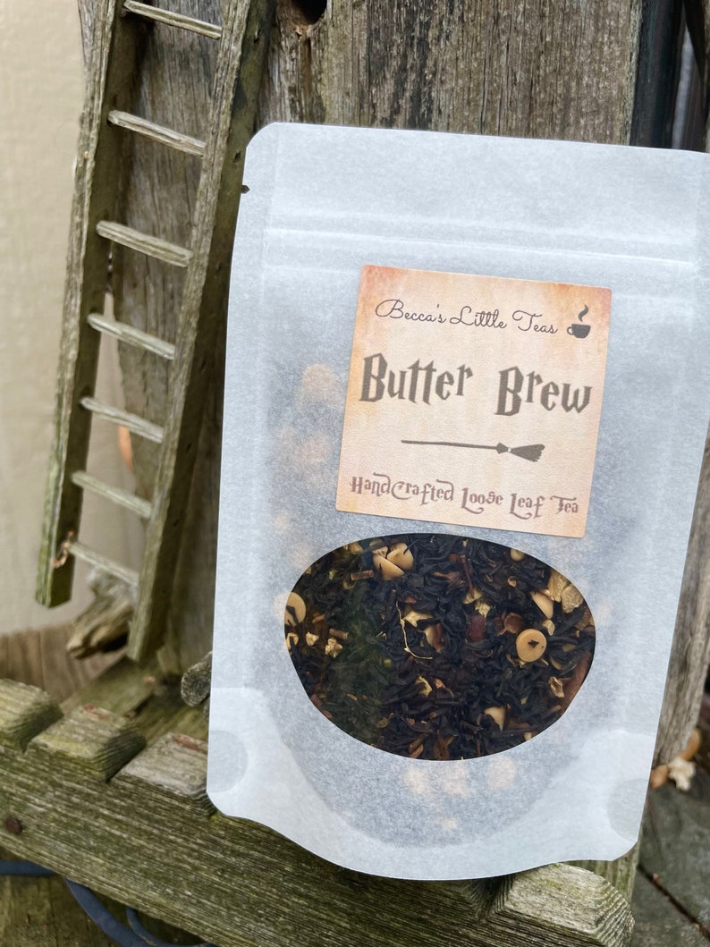 May include: A white resealable pouch with a clear window showing loose leaf tea. The label reads "Becca's Little Teas" and "Butter Brew" with a hand-drawn illustration of a broom. The label also says "Handcrafted Loose Leaf Tea".