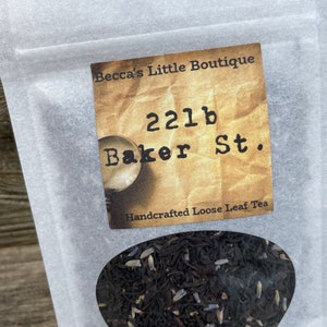 May include: A white paper bag with a label that reads "Becca's Little Boutique 221b Baker St. Handcrafted Loose Leaf Tea". The bag contains loose leaf tea with lavender buds.