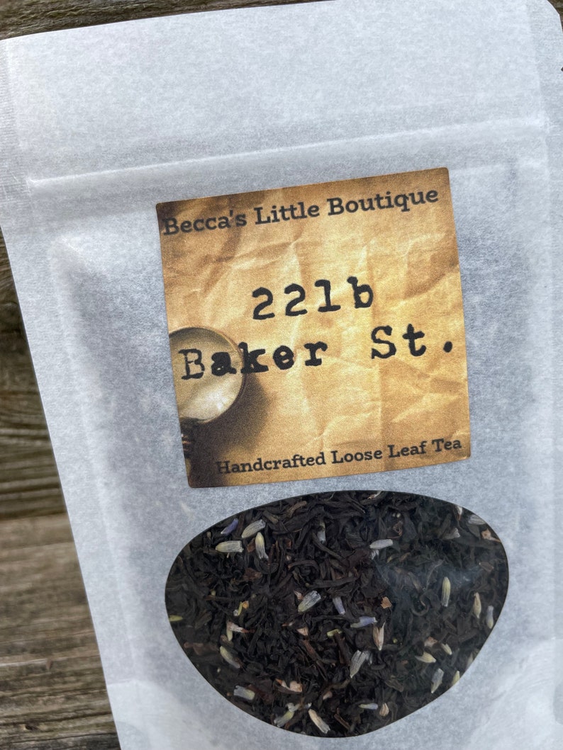 May include: A white paper bag with a label that reads "Becca's Little Boutique 221b Baker St. Handcrafted Loose Leaf Tea". The bag contains loose leaf tea with lavender buds.
