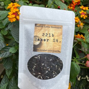 May include: A white paper bag with a label that reads "Becca's Little Boutique 221b Baker St. Handcrafted Loose Leaf Tea". The bag contains loose leaf tea.