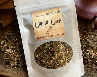 May include: A white paper pouch with a label that reads "Becca's Little Teas" and "Liquid Luck Handcrafted Loose Leaf Tea". The pouch is filled with loose leaf tea and is sitting on a wooden surface.