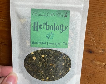 May include: A white paper pouch with a green label that says "Becca's Little Teas" and "Herbology Handcrafted Loose Leaf Tea". The pouch contains green tea leaves.