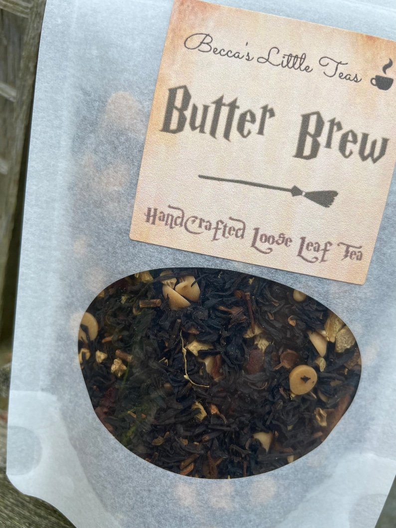 May include: A white paper bag with a clear window showing loose leaf tea. The bag has a label that reads "Becca's Little Teas" and "Butter Brew" in a dark brown font. The label also says "Handcrafted Loose Leaf Tea" in a smaller font.