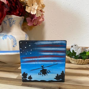 May include: A hand-painted wooden block featuring a night sky scene with a helicopter silhouette. The artwork includes a blue sky with red and white stripes, stars, and silhouetted trees. The block is painted with acrylic paints.