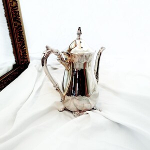 May include: A polished silver teapot featuring a curved handle with ornate detailing, a pointed lid with a decorative finial, and a slender spout. The teapot stands on small, sculpted feet and has a smooth, reflective surface throughout its body.