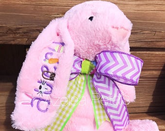 May include: A pink plush bunny with embroidered details on the ear. The ear features the name "Azalea" and a carrot and Easter egg design. A large bow with purple chevron and green gingham ribbons adorns the bunny's neck.