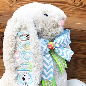 May include: A gray plush bunny with a blue and white chevron ribbon and a green and white gingham ribbon. The bunny has a personalized embroidered message that says "Happy Easter".