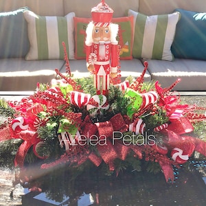 May include: A Christmas centerpiece featuring a nutcracker figurine, red and white candy canes, red glitter ribbon, and green foliage.