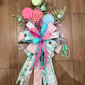 May include: A pink and blue Christmas tree topper with a bow, ribbons, and candy cane decorations. The ribbons feature a nutcracker pattern and the text "Joy to the World".