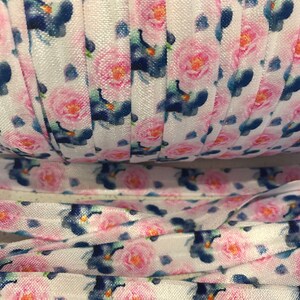 May include: White elastic with a floral print featuring pink roses and blue flowers. The elastic is rolled up and shows the pattern repeating.