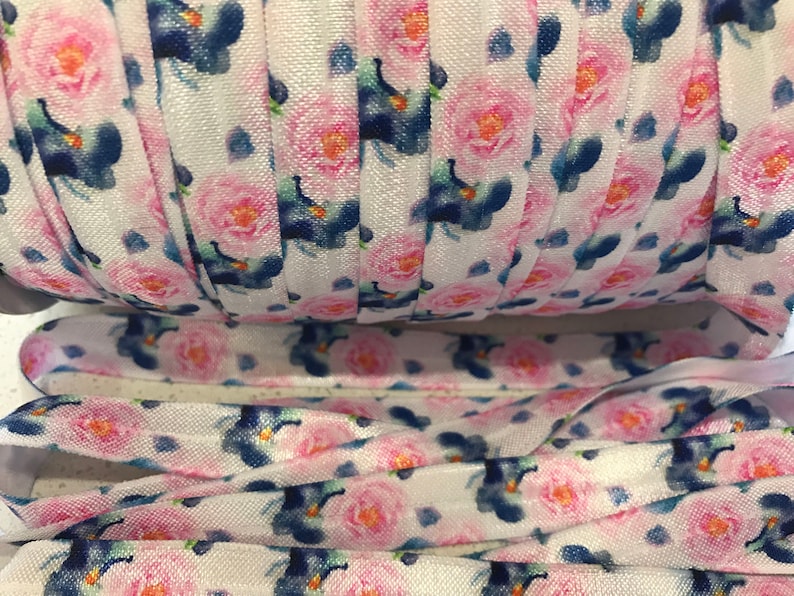 May include: White elastic with a floral print featuring pink roses and blue flowers. The elastic is rolled up and shows the pattern repeating.