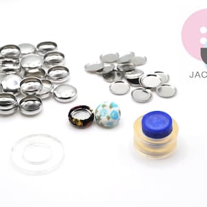 Puede incluir: Un juego de botones a presión de metal plateado, dos botones cubiertos de tela con motivos florales, un anillo de plástico transparente y una herramienta de presión de botones a presión de plástico azul y transparente. La herramienta está etiquetada como "JACKOBINDI".