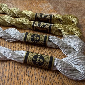 Op de afbeelding: Close-up van gouden en zilveren metallic borduurgaren strengen. Het garen is gelabeld met zwarte en gouden banden met de tekst "D.M.C. Metallic Pearl". De strengen zijn gerangschikt op een houten oppervlak.