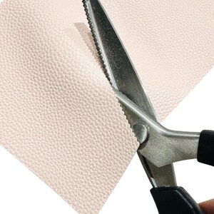 May include: Close-up of pink leather being cut with silver scissors. The scissors have black handles and are cutting a zig-zag pattern into the leather. The leather has a textured surface.