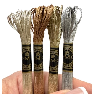 May include: Four spools of metallic embroidery thread in silver, gold, and bronze hues. Each spool features a black and gold label with the text "MOULINE M.C." and the measurement "m (8.7 yd)". The thread is held in a hand.