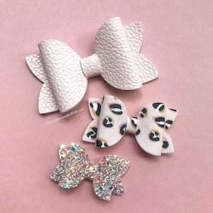 May include: Three hair bows of varying textures and patterns. The top bow is solid light pink with a pebbled texture. The middle bow features a black, gold, and white leopard print pattern. The bottom bow is covered in multi-coloured glitter.