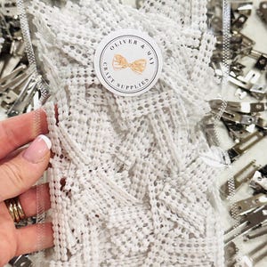 May include: A clear plastic bag filled with white, woven craft supplies. The bag has a circular sticker that reads "OLIVER & MAY CRAFT SUPPLIES" with a bow graphic. Silver metal clips are visible around the bag.