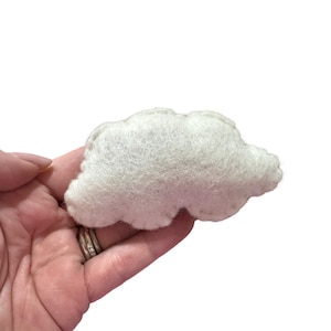 Może przedstawiać: Mała, biała chmurka z filcu. Chmurka jest puszysta i ma zaokrąglony kształt. Jest trzymana w dłoni osoby na białym tle. Jest to przedmiot rzemieślniczy.