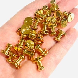 May include: A close-up of a hand holding a pile of gold-coloured metal fasteners. The fasteners have a round head and a threaded shank.