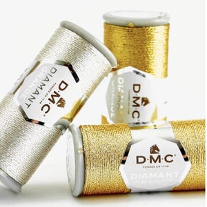Pode incluir: Três carretéis de linha de bordar metálica D.M.C. Diamant Grande. Um carretel é prateado e dois carretéis são dourados. Cada carretel apresenta um rótulo branco com o logótipo D.M.C., o texto Diamant Grande e a frase Fondée en 1746.