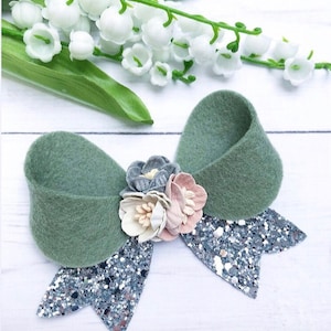 Puede incluir: Un lazo de fieltro verde con detalles de purpurina plateada y un grupo de tres flores de tela en tonos grises, rosa y blanco. El lazo está sobre una superficie blanca.
