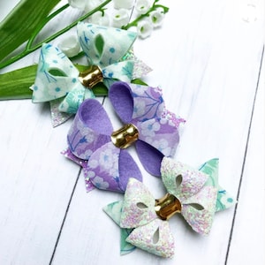 Puede incluir: Tres lazos para el pelo hechos a mano en una variedad de colores y patrones. Los lazos están hechos con una tela floral azul claro, una tela floral morada y una tela de purpurina blanca. Cada lazo tiene un centro dorado.