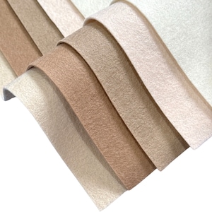 Puede incluir: Un conjunto de seis muestras de tela de fieltro en varios tonos de beige y marrón. Las muestras están dispuestas en forma de abanico, con el tono más claro en la parte superior y el tono más oscuro en la parte inferior.