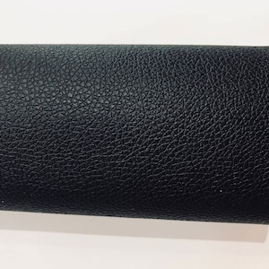 May include: A rolled piece of black faux leather with a textured, pebbled surface. The material is tightly rolled, revealing the underside and a glimpse of the white background. The leather appears to be durable and suitable for various crafting projects.