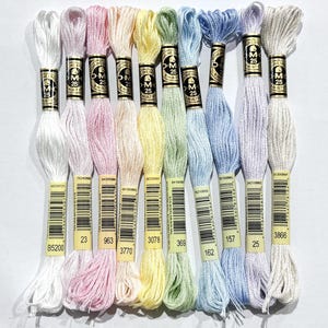 May include: A selection of DMC embroidery floss skeins in pastel shades. The colours include white, pink, yellow, green, blue, and lavender. Each skein is labelled with a number and colour code, ideal for cross-stitch and embroidery.