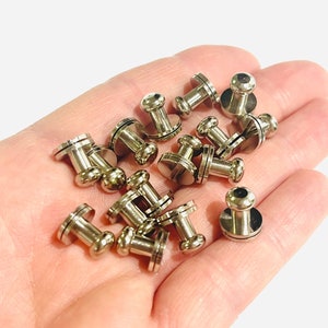 May include: A close-up of a pile of silver metal snap fasteners. The fasteners have a round head and a flat base.