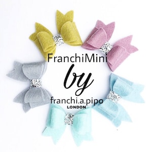 Puede incluir: Cinco lazos para el pelo de fieltro en diferentes colores: amarillo, gris, rosa, azul claro y turquesa. Cada lazo tiene un centro de purpurina plateada. Los lazos están dispuestos en un círculo con el texto "FranchiMini by franchi.a.pipo LONDON" en el centro.