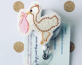 May include: A white glitter stork with a pink baby bag, holding a retractable badge holder with a butterfly design. The badge holder is attached to a white card with the text "LaAri's Custom Made Accessories" and "www.etsy.com/shop/" printed on it.