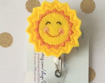 May include: A yellow felt sun with a smiling face and rosy cheeks is attached to a clear plastic badge holder. The badge holder has a metal clip and a spring-loaded retractable reel. The sun is attached to a white card with the text ".etsy.com/shop/l... email: droseman337@hotm... 10 to save 10% on your next order" printed on it.